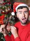Man in Santa hat holding open, empty wallet with Christmas tree in background.