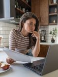 Woman with paper in one hand studying laptop.