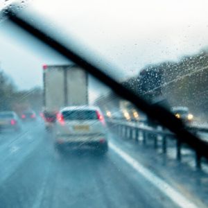 Car driving in rain from driver's POV with windshield wiper working.