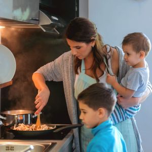 Mom holding baby while cooking dinner with child by her side.