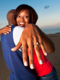 Engaged couple hugging in background; girl showing diamond ring on hand in foreground.